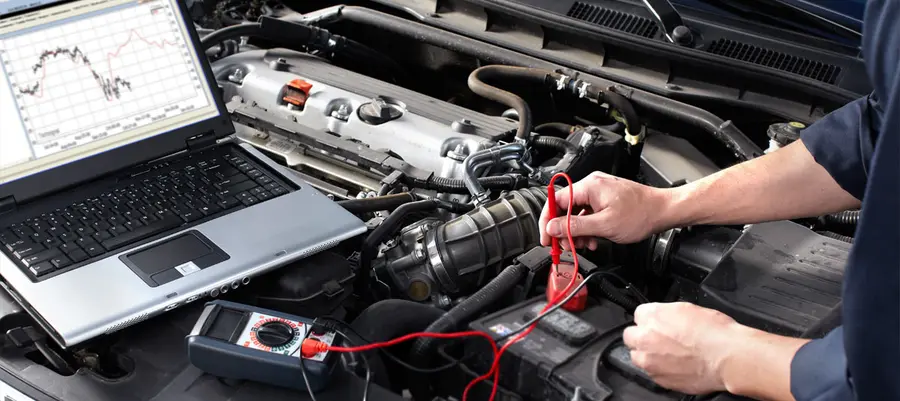 A mechanic checking a car engine bay with a meter, with a laptop resting beside the engine